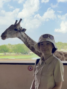 Iris, wearing a hat, standing in front of a giraffe. 