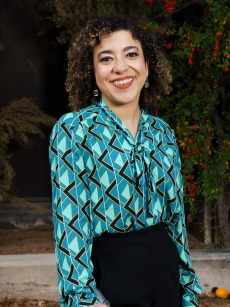Photograph of Dr. Kimberly Hassel. She is an Afro-Latina with short curly hair. She is wearing a teal shirt with abstract designs and black pants. She is smiling and looking at the camera.